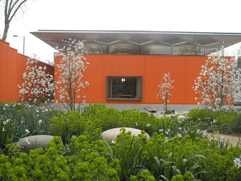 A modern looking orange building with silver roof. There is a nice garden in front of it.