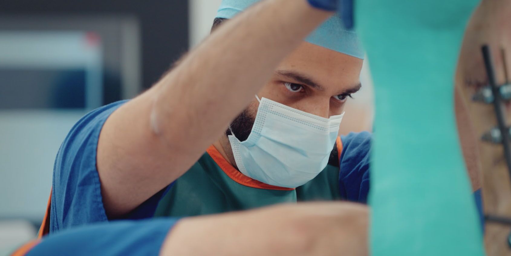 A surgeon wears blue scrubs and a face mask while he operates on a patient